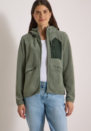 Kvinde iført en grøn fleecejakke med lynlås på brystet og siderne over en hvid skjorte og blå jeans, står mod en ensfarvet baggrund.