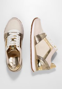 Sneakers w kolorze kremowym i złotym, z teksturowanymi wykończeniami, wiązaniem na sznurówki oraz metalicznymi akcentami po bokach i na pięcie.