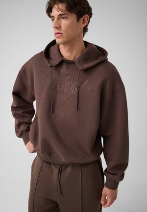 Jeune homme portant un sweat à capuche marron et un pantalon assorti, regardant sur le côté avec une main dans la poche, sur un fond uni.