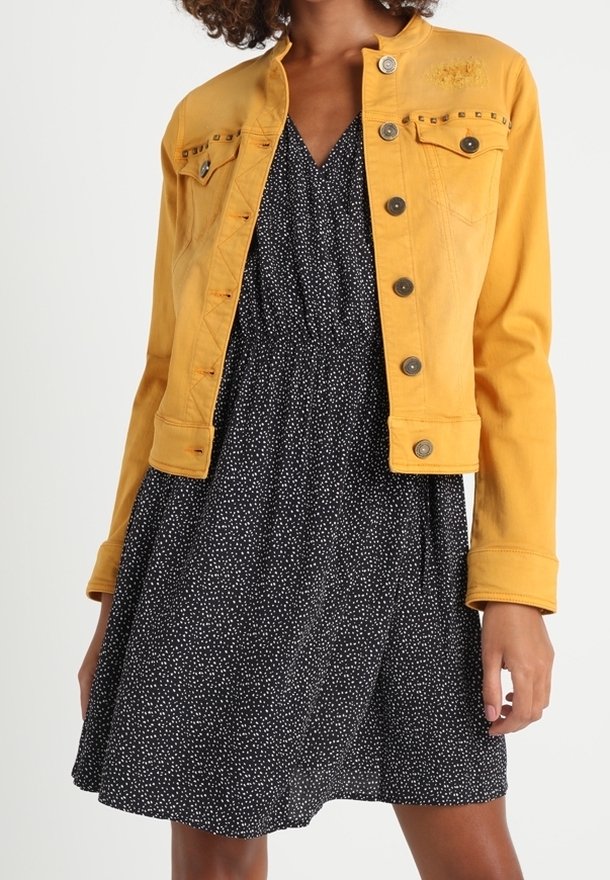 Žena oblečená v hořčicově žluté bundě na knoflíky přes černé šaty s bílými puntíky, stojící na jednobarevném pozadí.