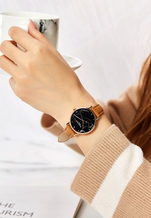 Montre noire ronde avec un design marbré, lunette dorée et bracelet en cuir beige. Main reposant sur une tasse au motif marbré. Pull neutre.