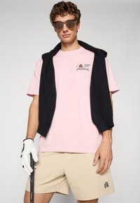 Lys rosa t-skjorte med grafikk, svart genser knyttet rundt skuldrene, beige shorts, hvit golfhanske, holder en golfklubbe. Brune solbriller.