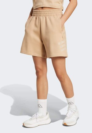 Pantaloni corti beige in morbido tessuto con vita elasticizzata, tasche laterali e tre strisce chiare sulla gamba sinistra. Indossati con sneakers bianche e calze.