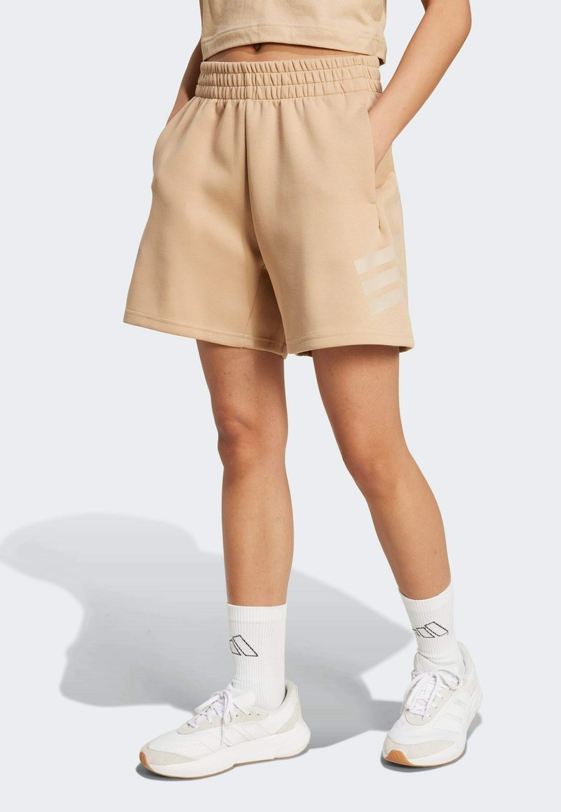 Pantalones cortos de tela suave en beige con cinturilla elástica, bolsillos laterales y tres rayas claras en la pierna izquierda. Llevados con zapatillas blancas y calcetines.