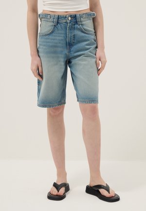 Persona che indossa pantaloncini di jeans blu slavato di media lunghezza, top bianco corto e infradito platform nere, in piedi davanti a uno sfondo semplice.