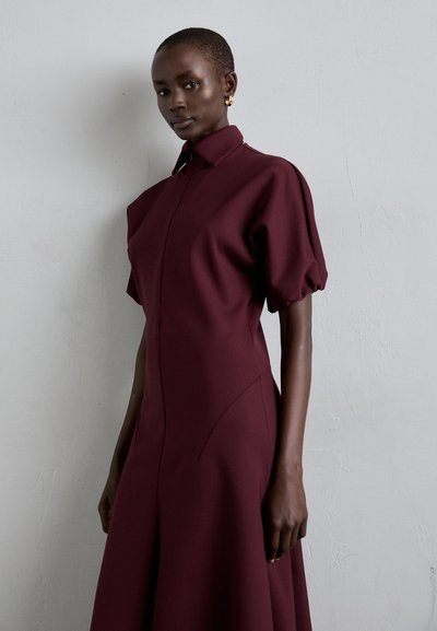 Rochie burgundy cu un corset ajustat, maneci bufante și un guler înalt. Material neted cu detalii pe cusăturile laterale; fustă de lungime până la genunchi.