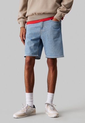 Shorts in denim azzurro con vestibilità comoda, dotati di tasche laterali e orlo sotto il ginocchio. Abbinati a una felpa beige e sneakers chiare.