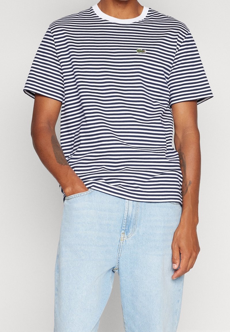 Man wearing navy and white striped short-sleeve T-shirt with small green crocodile logo and light blue denim jeans, one hand in pocket.