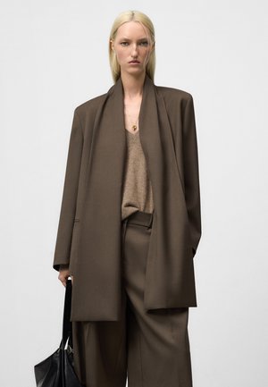 Vrouw met stijlvol blond haar in een oversized bruin pak, beige V-hals trui, gouden ketting, met een zwarte leren tas, op een neutrale achtergrond.