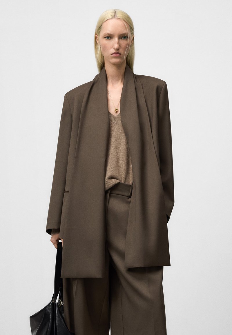 Frau mit glattem, blondem Haar, die einen übergroßen braunen Anzug, einen beigen V-Ausschnitt-Pullover und eine goldene Halskette trägt. Sie hält eine schwarze Ledertasche und steht vor einem neutralen Hintergrund.