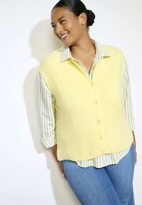 Geltonas be rankovių megztinis su mygtukais, pagamintas iš tekstūruotos medžiagos, uždėtas ant mėlynos ir baltos juostuotos marškinėlių, dėvimas su džinsinio audinio kelnėmis.