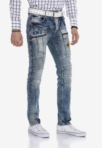 Blaue Jeans aus Denim mit mehreren Taschen, abgenutzter Textur, Patches und gestickten Details. Getragen mit einem weißen Nieten-Gürtel und Sneakers.