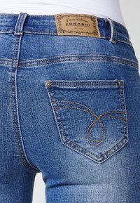 Blå denimjeans med en brun läderlapp med logotyp, bakfickor med kontrasterande sömmar och en texturerad tygfinish.