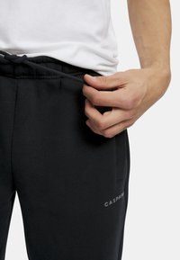 Svarta sweatpants med en mjuk textur, featuring en elastisk midja och snörning. Varumärket "CASPARI" visas i vita bokstäver.