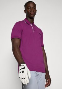 Vijolična polo majica s klasičnim ovratnikom, ki ima bel trak in črne obrobe. Bele golf rokavice dopolnjujejo izgled z moderno in čisto obliko.