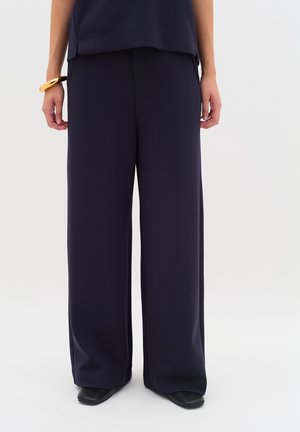 Person wearing loose, wide-leg navy pants, black shoes, a matching navy top, and a large gold bracelet on left wrist.