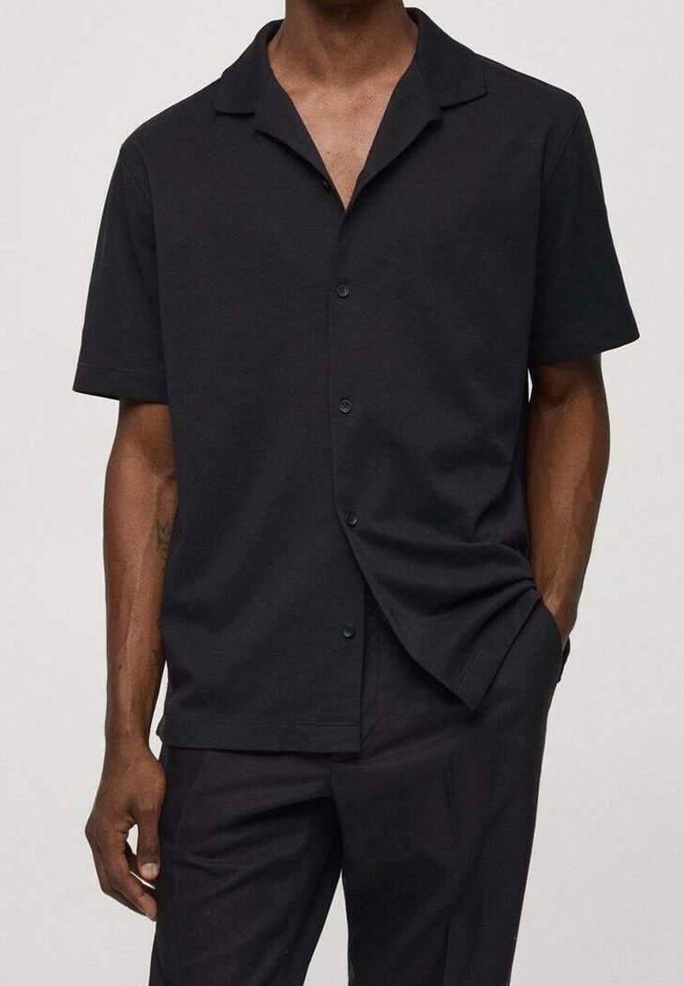Homme portant une chemise noire à manches courtes à boutons avec col, associée à un pantalon noir, une main dans la poche, fond neutre.