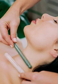 Dwa narzędzia do masażu, jedno zielone, a drugie białe, są umieszczone wzdłuż szyi osoby, podczas gdy jedna ręka trzyma zielone narzędzie. Widoczny neutralny odcień skóry.