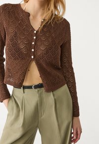 Bruine gebreide cardigan met een ruitpatroon, voorzien van een ronde halslijn en een witte knoopsluiting. Gepaarde met hooggetailleerde beige broeken.