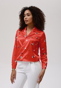 Blusa di raso rosso con motivi floreali in rosa, nero e giallo. Presenta un colletto, maniche lunghe e un orlo corto. Abbinata a pantaloni bianchi.