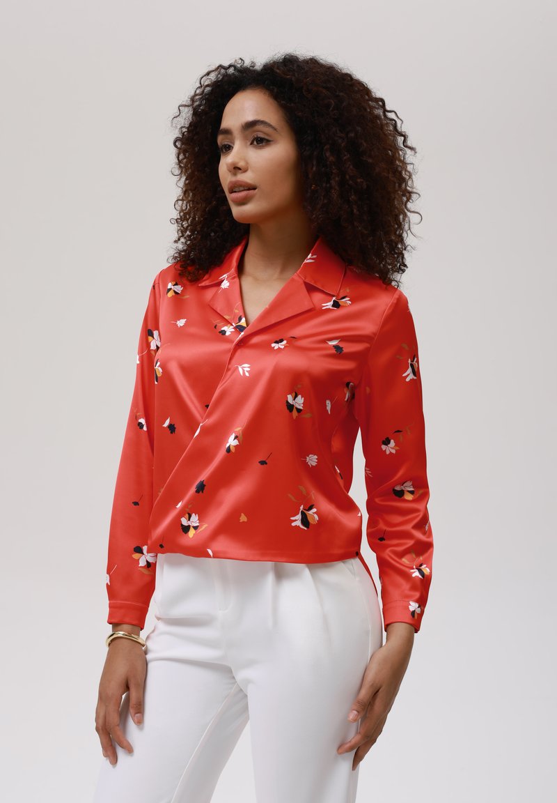 Blusa di raso rosso con motivi floreali in rosa, nero e giallo. Presenta un colletto, maniche lunghe e un orlo corto. Abbinata a pantaloni bianchi.
