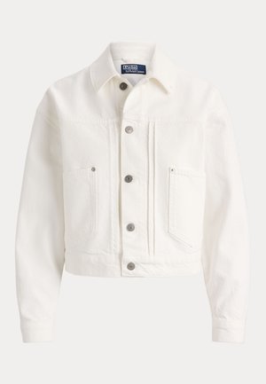 Frau mit geflochtenem Haar, die eine weiße, kurze Jacke, ein weißes Oberteil, weite marineblaue Hosen und schwarze offene High Heels trägt, steht vor einem neutralen Hintergrund.