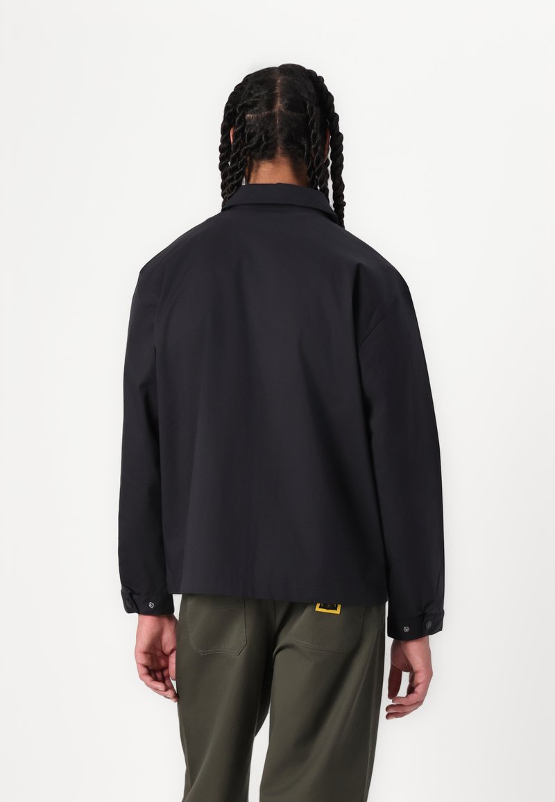Veste noire légère avec un col, des manches longues et une coupe décontractée. Comprend des poignets boutonnés et un accent logo près de l'ourlet.