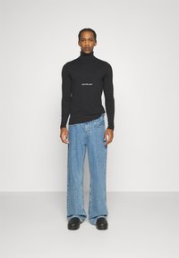Maglione a collo alto nero con il logo bianco "Calvin Klein Jeans", abbinato a jeans in denim azzurro chiaro a gamba larga e calzature nere.