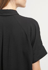 Camicia nera con colletto e maniche corte arrotolate. Realizzata in un tessuto liscio, presenta una leggera lucentezza e una trama sottile nel materiale.