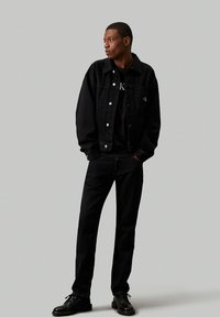 Chaqueta de mezclilla negra con botones frontales y bolsillos, usada sobre una camisa negra y combinada con jeans y zapatos negros, frente a un fondo liso.