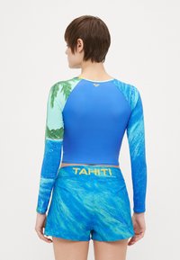 Långärmad baddräkt i blått med grön palmtamönster; matchad med blå shorts som har "TAHITI" i gult på midjebandet.
