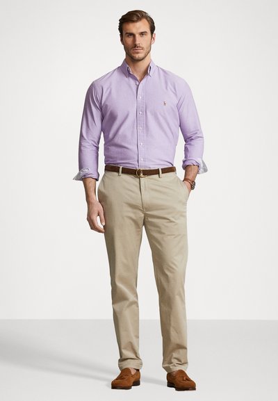 Ανοιχτό μωβ πουκάμισο με γυρισμένα μανίκια, σε συνδυασμό με ανοιχτό μπεζ παντελόνι και καφέ loafers, που στέκεται σε ένα απλό φόντο.