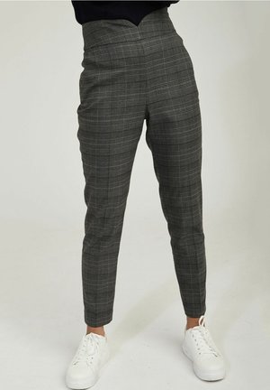 Person iført grå ternede højgyldne smalle bukser kombineret med hvide sneakers, stående foran en ensfarvet lys baggrund.