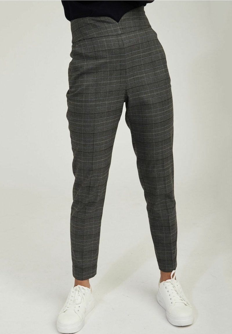 Personne portant un pantalon à carreaux gris taille haute et fuselé, associé à des baskets blanches, debout devant un fond clair uni.