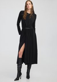 Czarna sukienka z długim rękawem i skręconym detalem w talii, z bocznym rozcięciem i srebrnym akcentem. Połączona z czarnymi kozakami na wysokim obcasie do kolan.