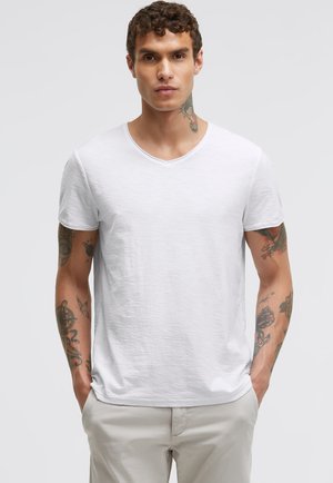 Mann mit lockigem Haar und mehreren Tätowierungen an Armen und Hals steht mit den Händen in den Taschen und trägt ein schlichtes weißes T-Shirt mit kurzen Ärmeln und V-Ausschnitt.