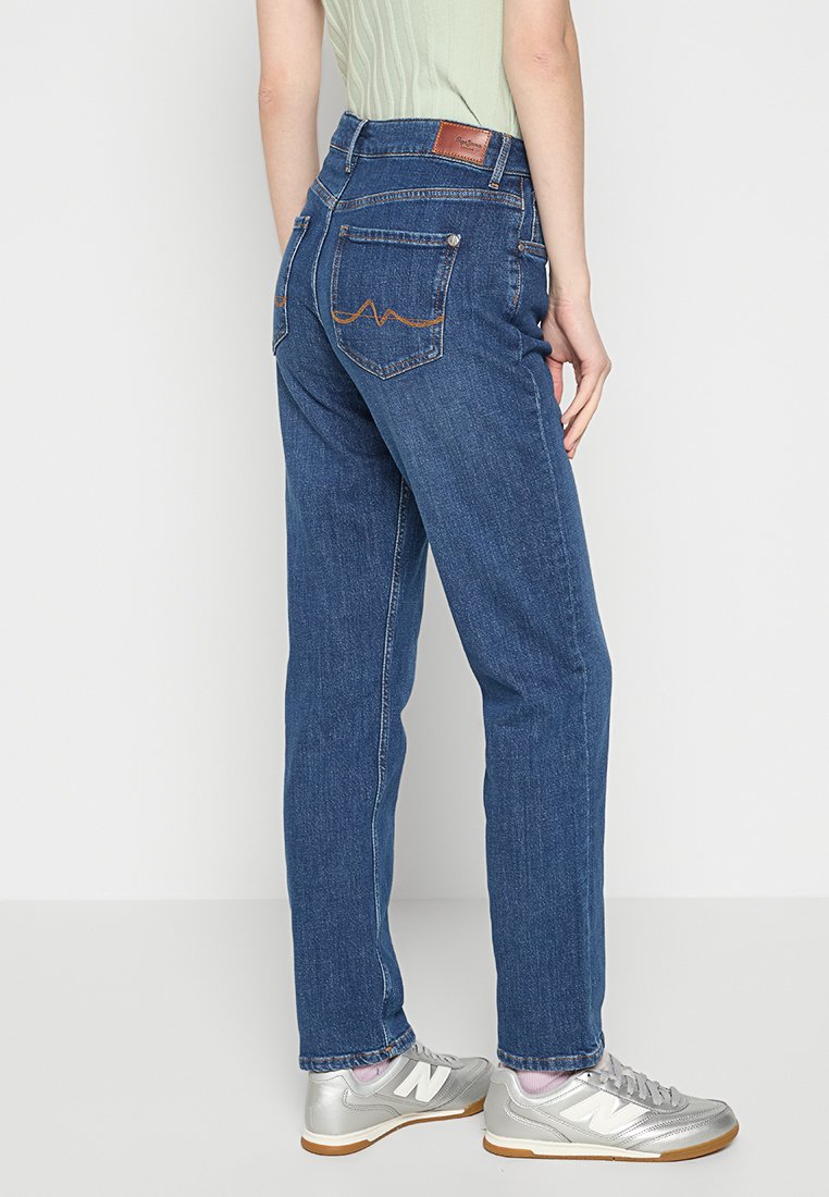 Person, der hoch taillierte blaue Jeans mit bestickten Gesäßtaschen, ein hellgrünes geripptes Oberteil und silberne Sneaker mit Gummisohlen trägt.