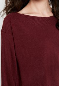 Maglione a maglia borgogna con scollatura larga a barca, spalle cadenti e una consistenza morbida, con finiture a coste sul collo e sui polsini.