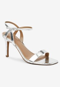 Sandal in metallo argentato con cinturini, punta quadrata, tacco sottile e cinturino alla caviglia regolabile. Presenta una superficie lucida e texture.