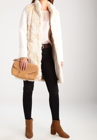 Cremefarbene Kunstfelljacke mit langen Ärmeln, kombiniert mit einem weißen Oberteil, schwarzen engen Jeans und braunen Stiefeletten. Hält eine beige Clutch-Tasche.