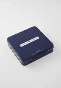 Sötétkék fémtartó lekerekített sarkakkal, fehér "TOMMY HILFIGER" logóval és címkével, amely a zoknik méreteit és mennyiségeit tartalmazza.