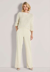 Vrouw in een off-white, gestructureerd gebreid sweater en wijde crèmekleurige corduroy broek staat tegen een effen neutrale achtergrond.