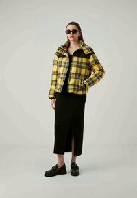 Mulher com óculos de sol pretos, casaco acolchoado amarelo em padrão xadrez, saia midi preta com fenda frontal e loafers pretos de sola grossa, posando contra um fundo simples.