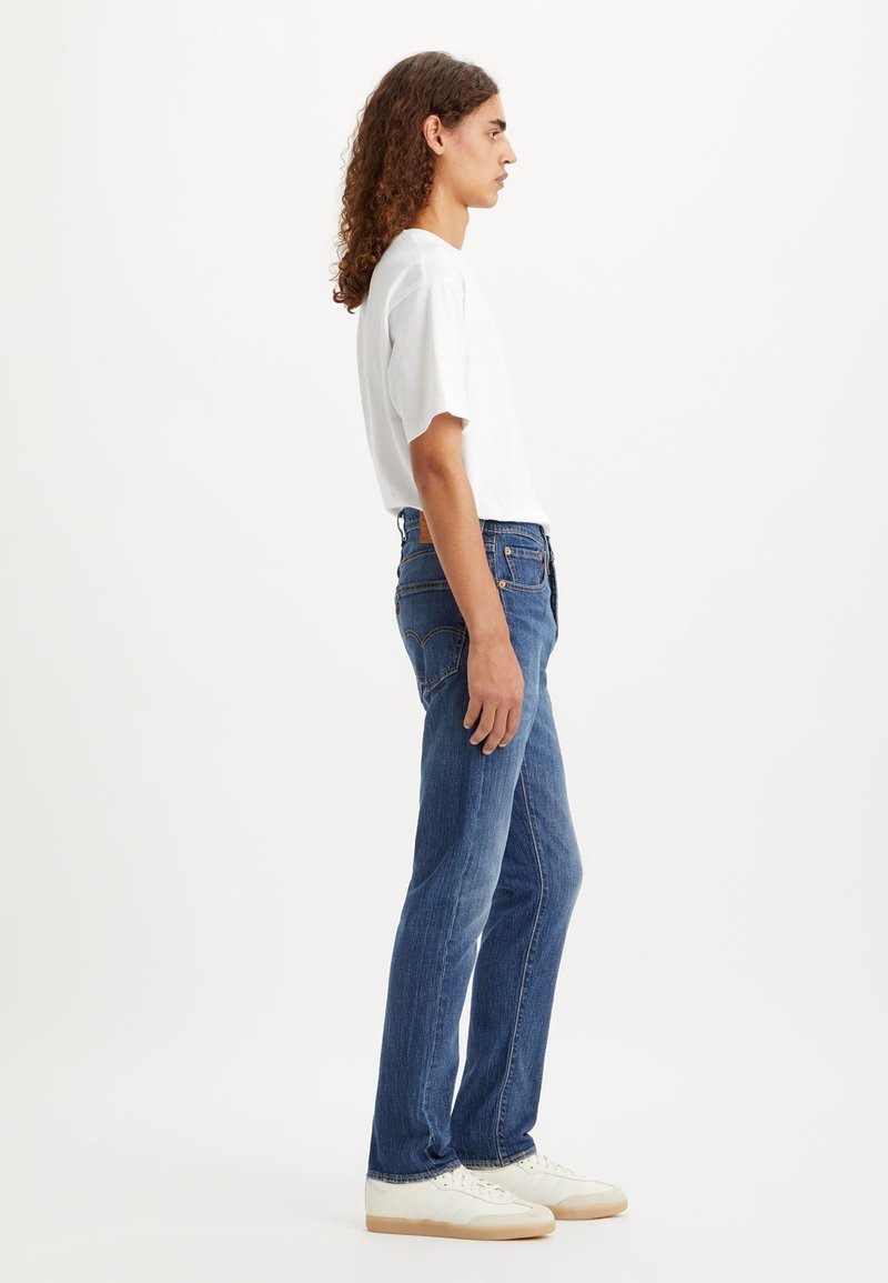 Blaue Jeans aus Denim mit geradem Bein, ausgestattet mit fünf Taschen und einem Lederschild auf der Rückseite. Kombiniert mit einem weißen T-Shirt und hellen Schuhen.