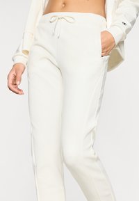Κρεμ φόρμες jogger από απαλό ύφασμα, με κορδόνι στη μέση, πλαϊνές τσέπες και λογότυπο στο ισχίο. Εφαρμοστή, κωνική γραμμή.