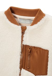 Veste en polaire couleur crème avec un col à côtes marron, fermeture éclair à l'avant et une poche zippée marron sur le côté gauche. Texture douce.