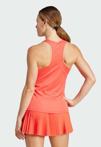 Haut de sport à manches courtes, de couleur corail vif, avec un design dos nageur ; associé à une jupe plissée corail. Tissu lisse, coupe ajustée.