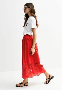 Gonna midi rossa in cotone con dettagli floreali a taglio sul bordo, abbinata a una t-shirt bianca e sandali neri, mostrata di lato.