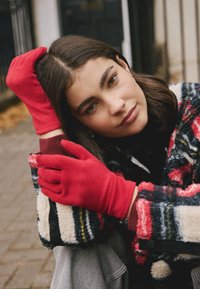 Gants en polaire rouge à coupe ajustée, assortis à un manteau en laine à motifs rouge, noir et jaune, porté sur un haut noir.