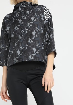 Vrouw die een pullover draagt met een zwart-grijs camouflagepatroon en driekwart mouwen, gecombineerd met zwarte sportleggings, tegen een witte achtergrond.